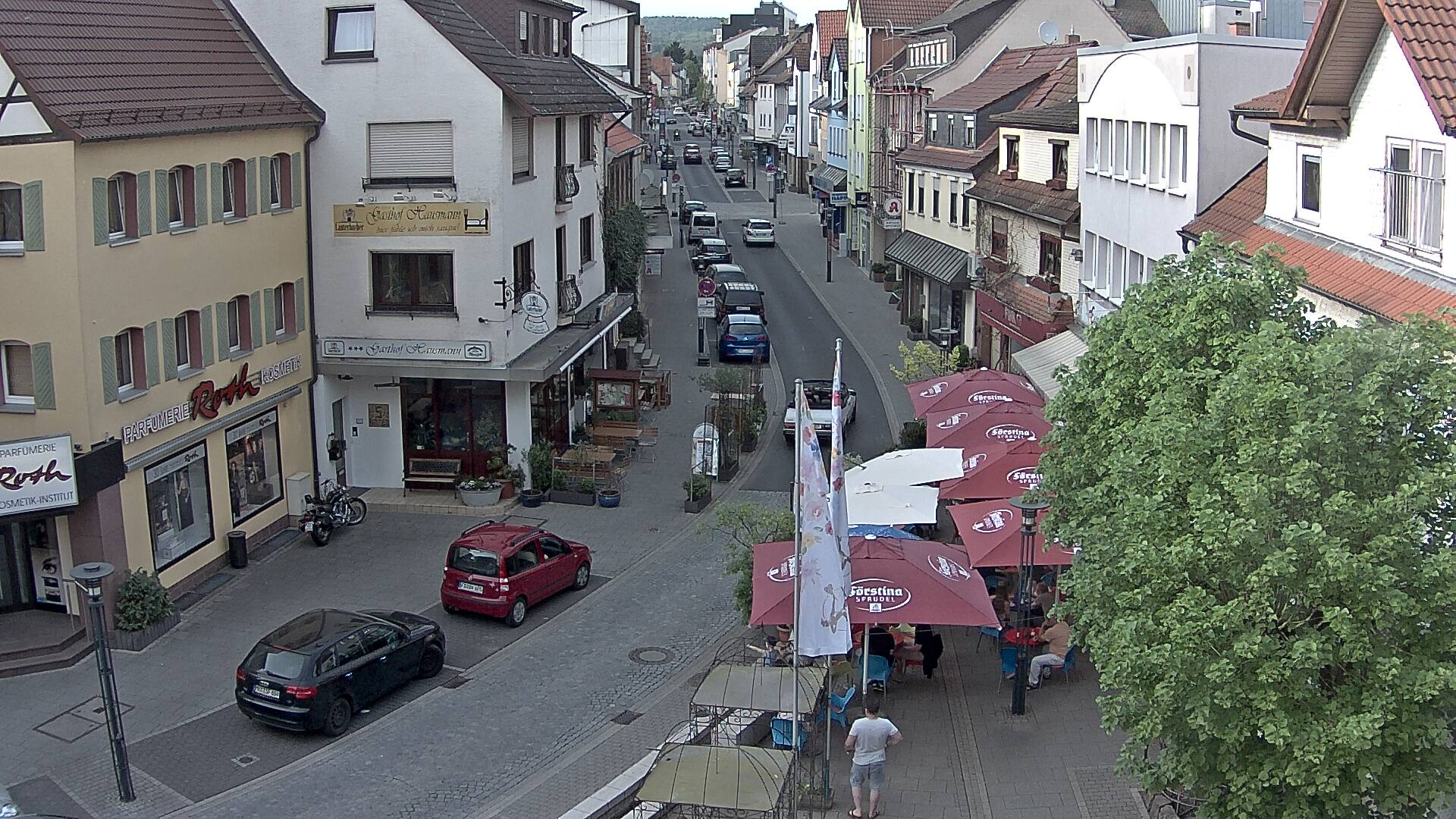 Blick von oben in eine belebte Straße mit breiten Gehwegen, rechts und links stehen Häuser. Vorn rechts ein Straßencafé mit Sonnenschirmen