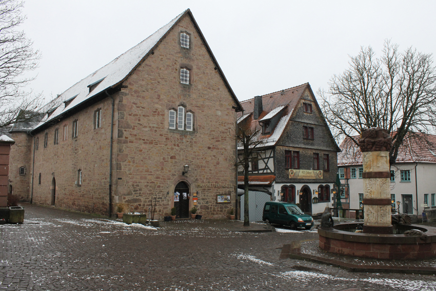 Blick auf einen menschenleeren Platz im Winter. In der Bildmitte die Giebelseiten von zwei historischen Gebäuden, rechts der Märchenbrunnen mit einer Säule.